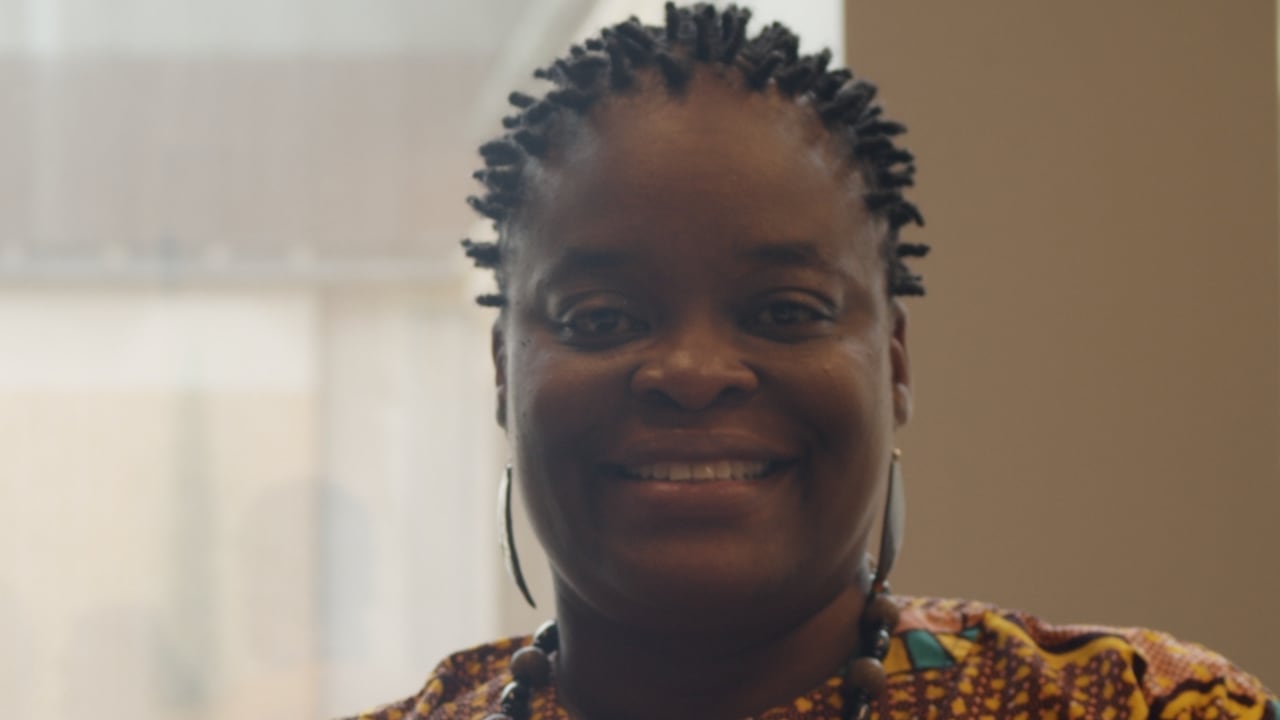 A Black woman wearing a colourful blouse smiles at the camera.