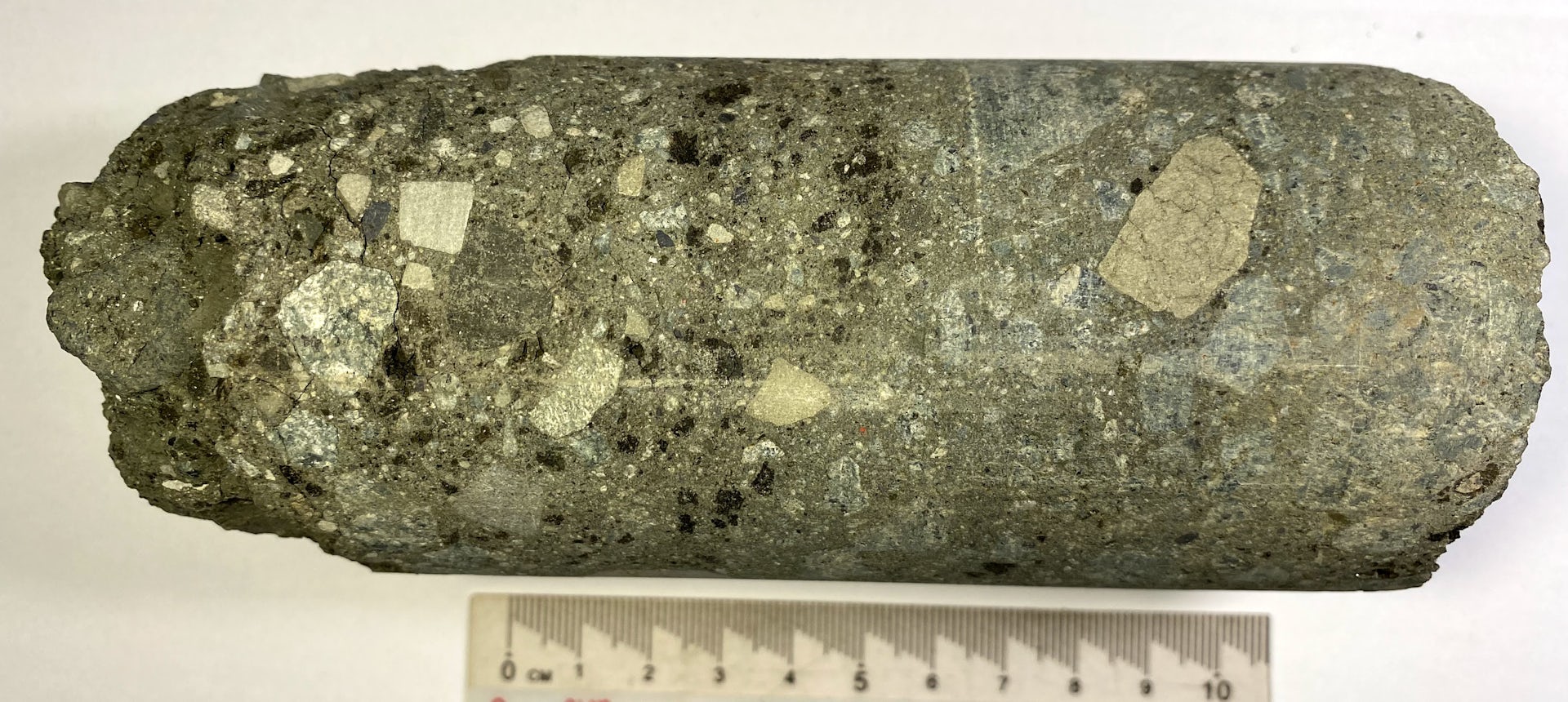 A cylindrical drill core sample approximately 10 cm long, composed of a dark grey-green fine-grained matrix containing numerous white to cream-colored angular clasts of varying sizes, scattered black mineral grains, and minor pyrite. A ruler in centimeters is shown below for scale. The texture is consistent with a volcaniclastic or impact breccia lithology.