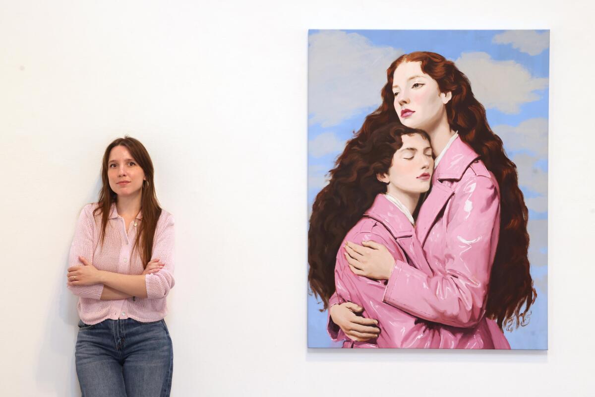 A woman stands next to a portrait of two women, wearing pink vinyl jackets, hugging.