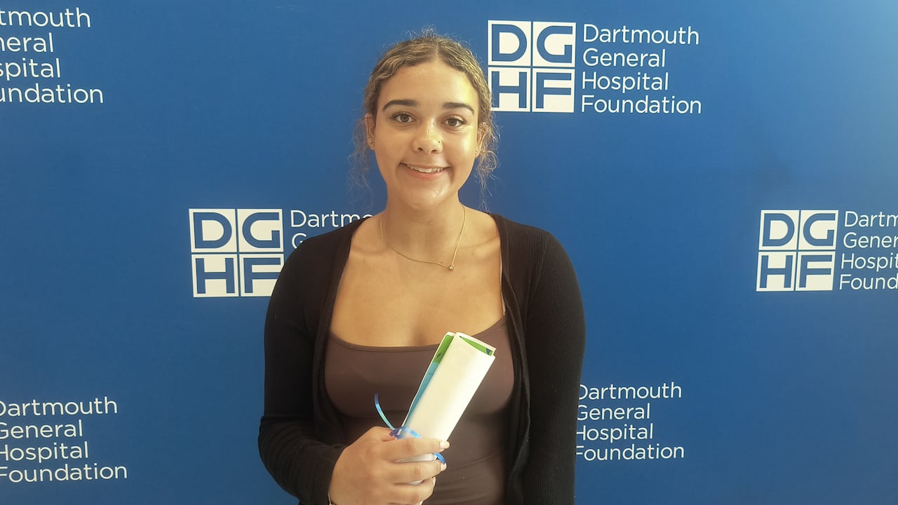 A girl smiles while holding a diploma. She is in front of a blue background.