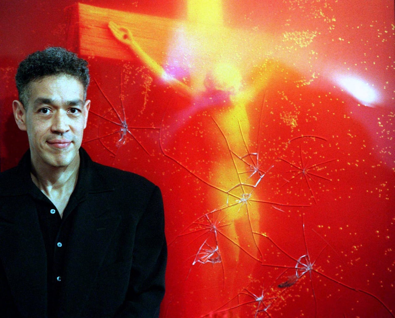 A man with a neutral expression stands next to a photograph he took, which shows a crucifix sitting in urine.
