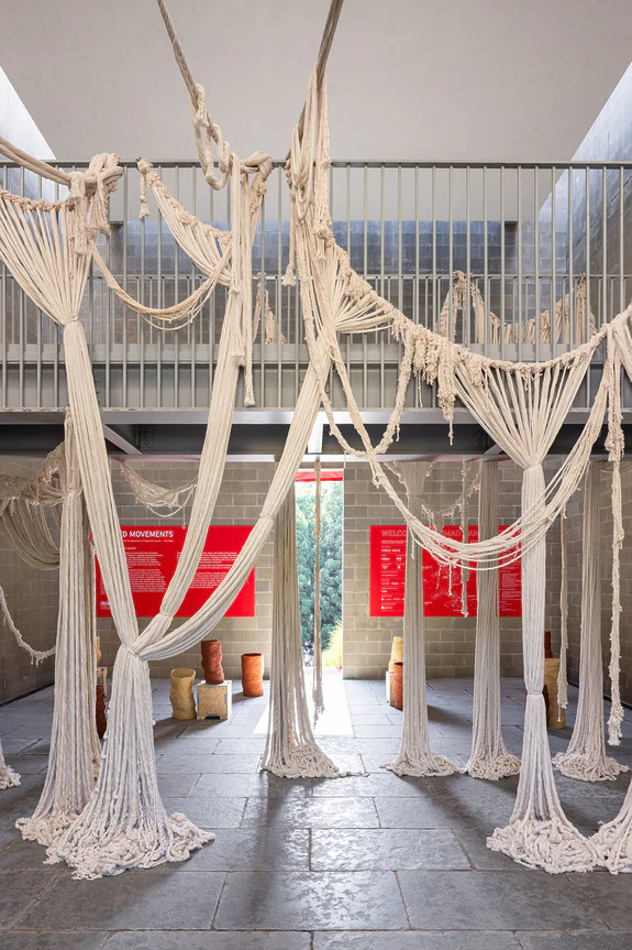 Textile art installation with draped fabric structures hanging from the ceiling in a spacious gallery setting.