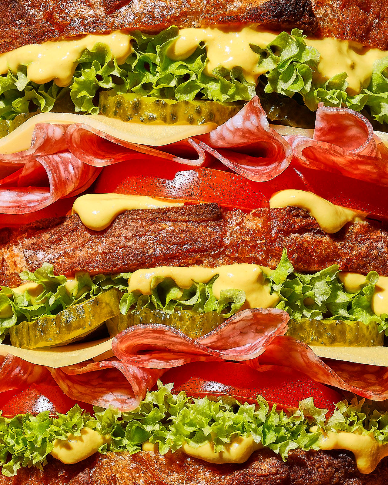 Close-up of a stacked sandwich with layers of lettuce, tomato, pickles, sliced salami, cheese, and mustard between slices of brown bread.