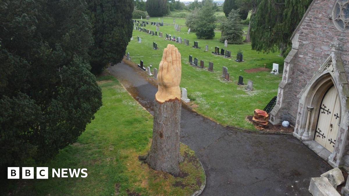 Aerial shot of a stump of a tree. It now has two hands praying at the top of it. It is surrounded by trees and in a cemetery with graveyards in the background.