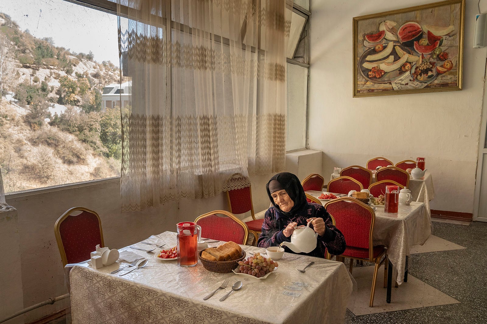 An elderly woman in a headscarf sits alone at a table in a sunlit restaurant, pouring tea. The table has bread, fruit, and red pitchers. Large windows reveal a rocky landscape outside. A fruit painting hangs on the wall.