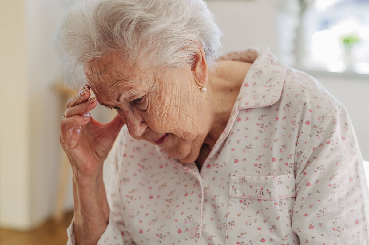 Elderly woman in pyjamas sitting on bed, holding her head.
