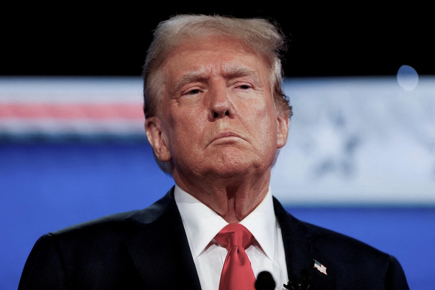 An older man — Donald Trump — wearing a red tie, white shirt and a black suit jacket with a serious expression.