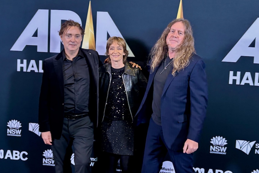 Two long-haired men flank a shorter and younger woman at an awards' night