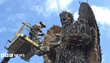 A man on a cherry picker with grey clothing and a helmet facing a large metal sculpture of an angel made from hundreds of knives