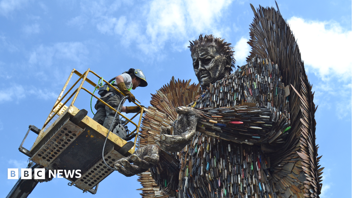 A man on a cherry picker with grey clothing and a helmet facing a large metal sculpture of an angel made from hundreds of knives