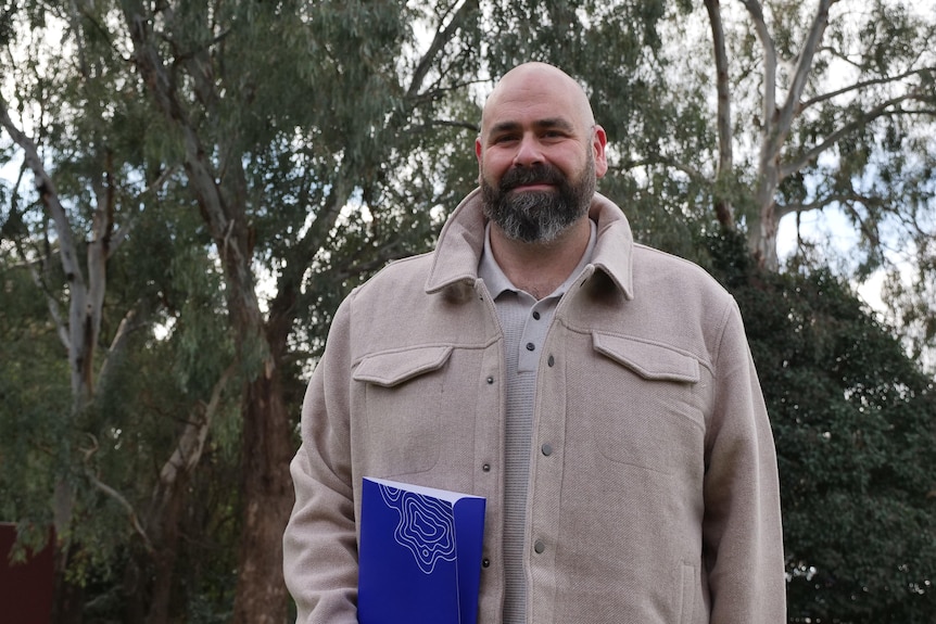 A man in a jacket holding a folder standing in front of trees