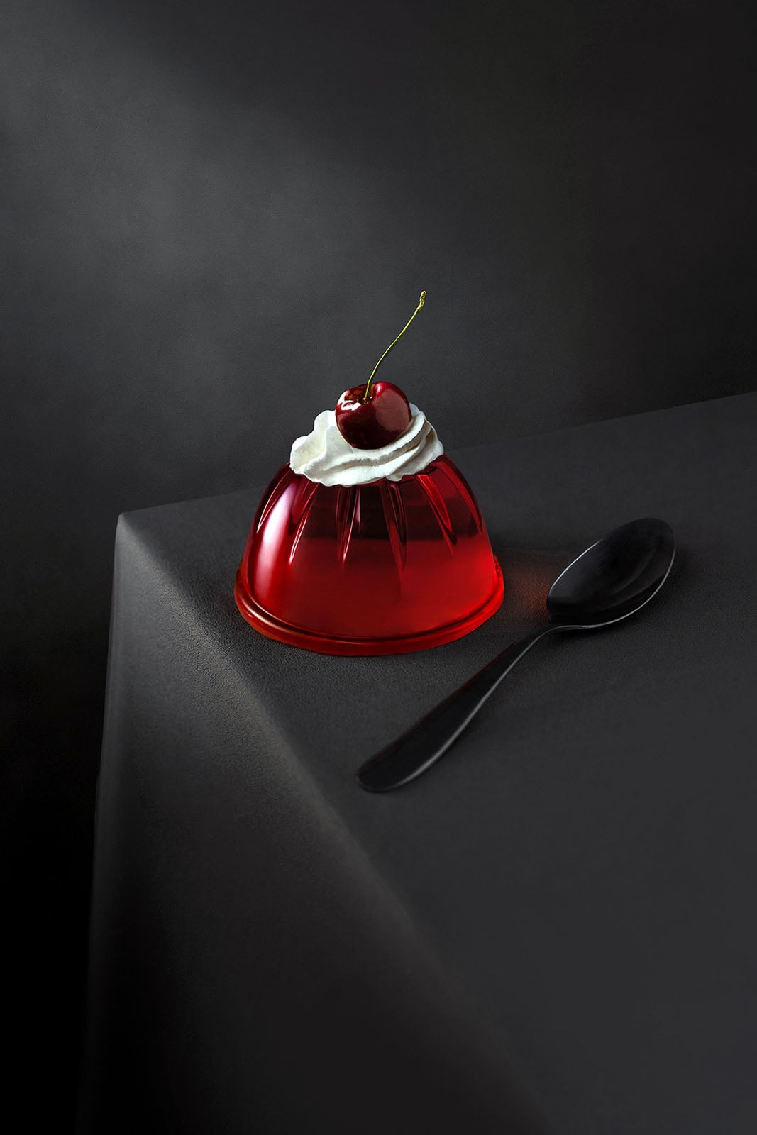 A red gelatin dessert topped with whipped cream and a cherry sits on a dark tablecloth next to a black spoon, against a dark background.
