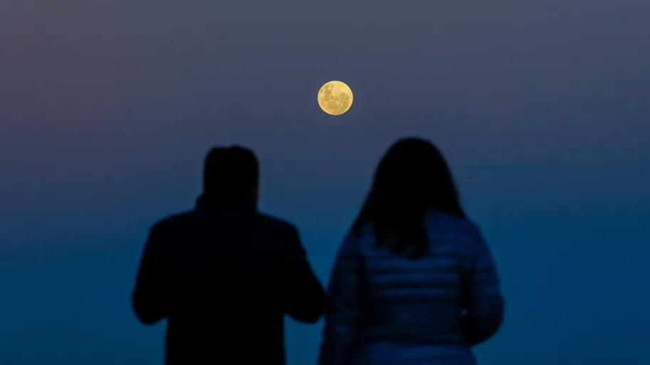 Two people sit with their backs facing the camera, watching the Strawberry Moon in the sky