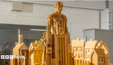 A model sculpture made of yellow wood depicts a miner rising out of some colliery buildings. He is wearing a miner's hat and lamp.