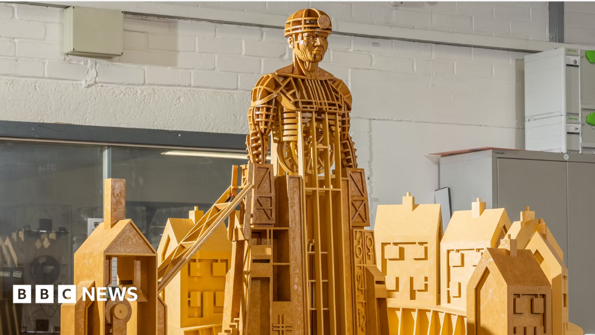 A model sculpture made of yellow wood depicts a miner rising out of some colliery buildings. He is wearing a miner's hat and lamp.