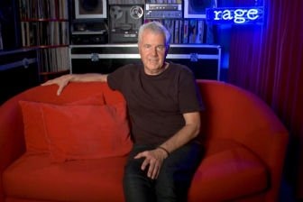 A man in a dark T-shirt sits on a red couch, looking at the camera.