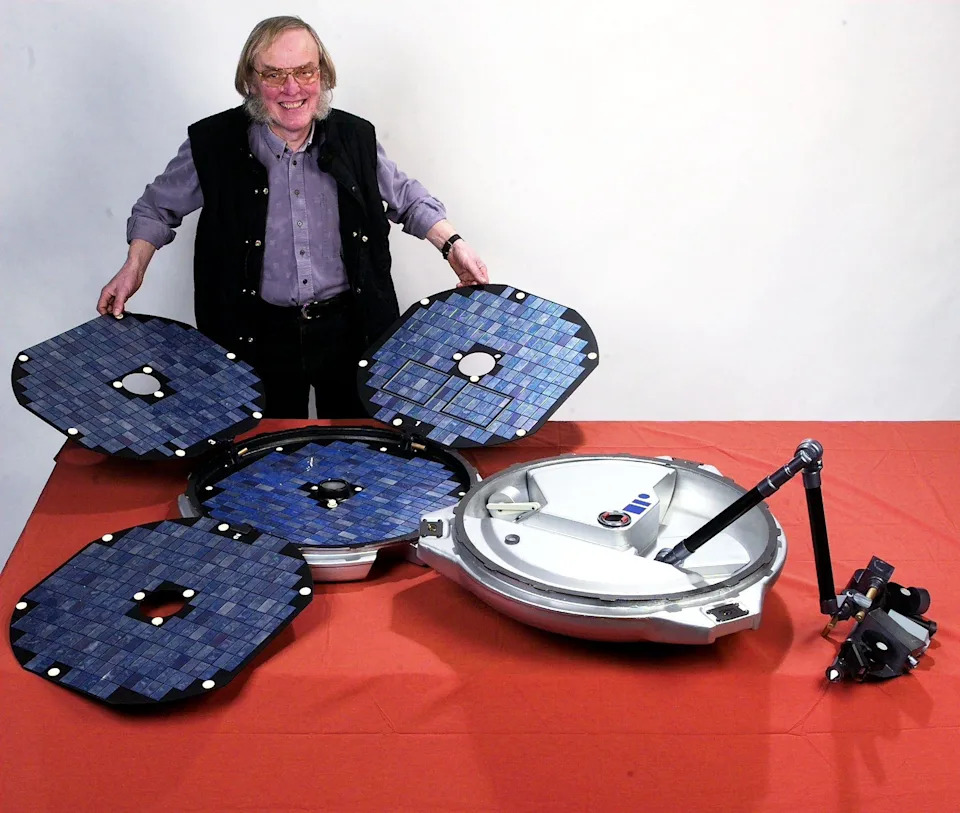 An older man with large sideburns is holding parts of a Mars lander, specifically solar panels