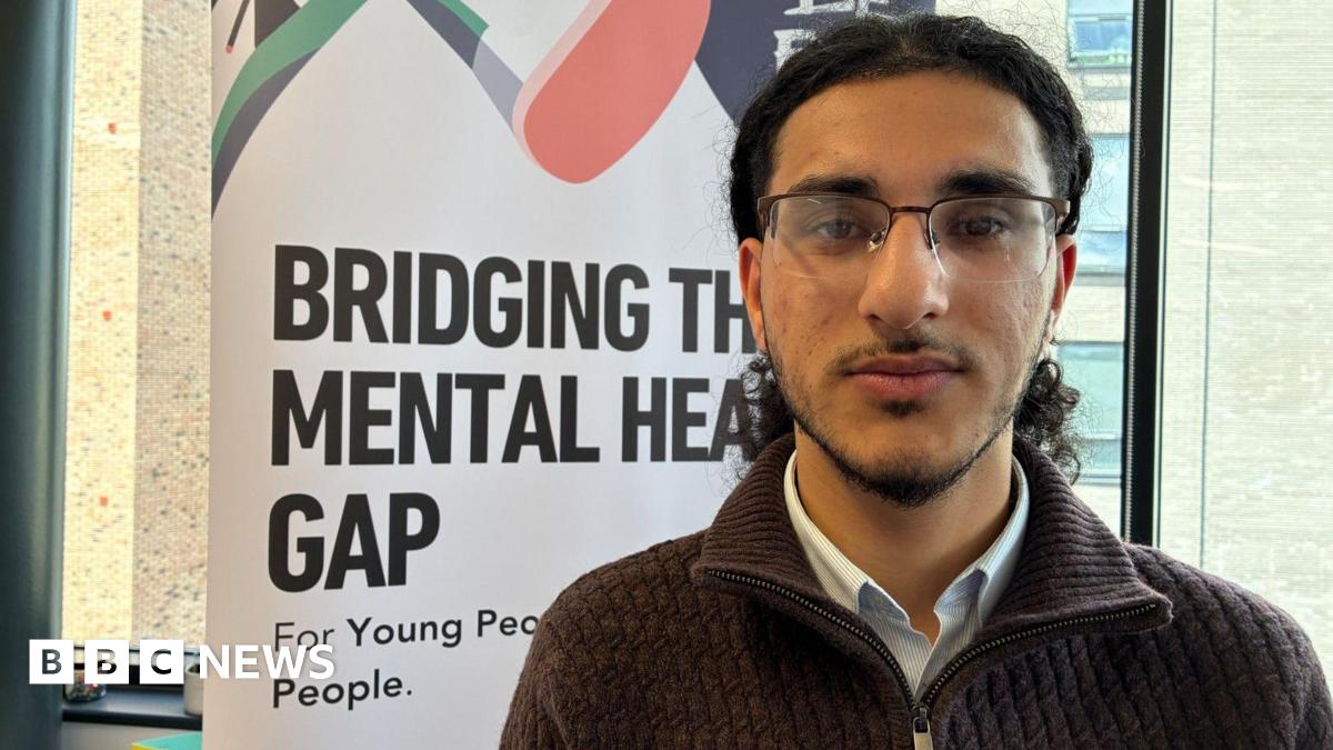 Rushid has long black hair and a beard and is wearing glasses, a blue and white shirt and a brown zipped top. He is standing in front of a white banner which says "bridging the mental health gap" in black letters.