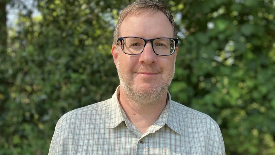 Man with short brown hair and moustache and beard and wearing glasses smiling at the camera. He is wearing a checked shirt that is Forestry Commission workgear. 
