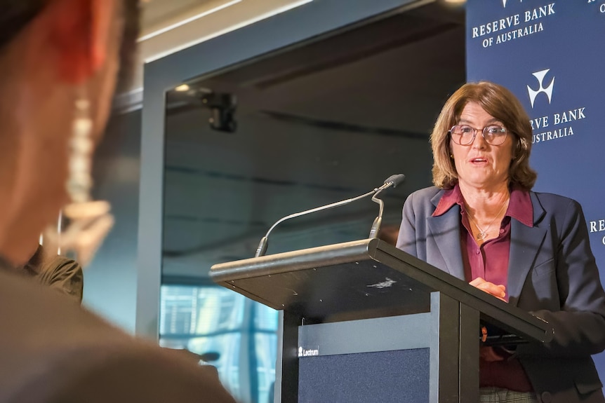A dark-haired woman wearing glasses and a blazer leans on a lectern in front of an RBA-branded backdrop.