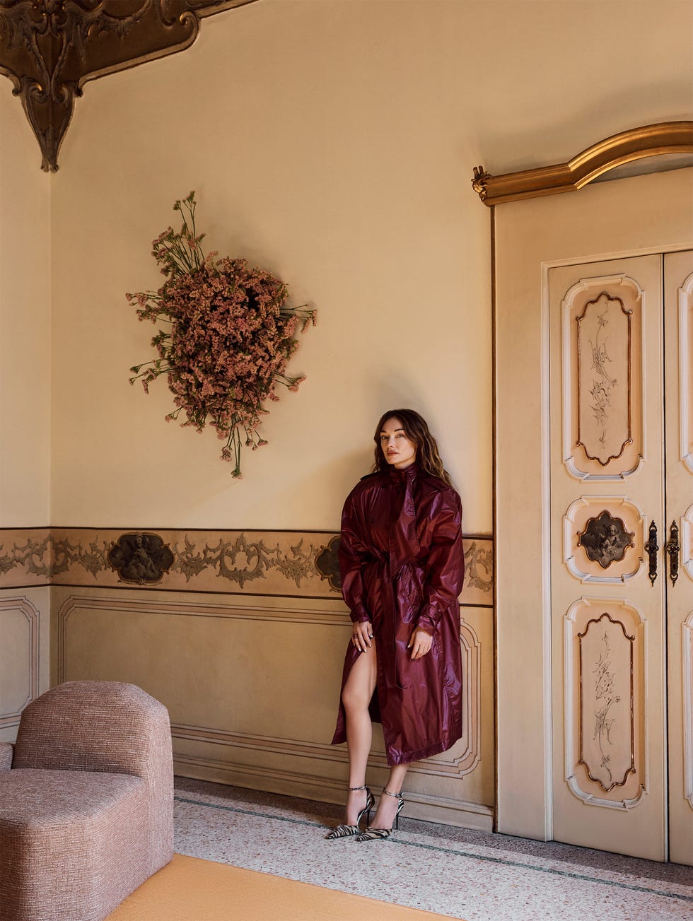 Person in maroon coat standing in ornate room with floral wall decoration.