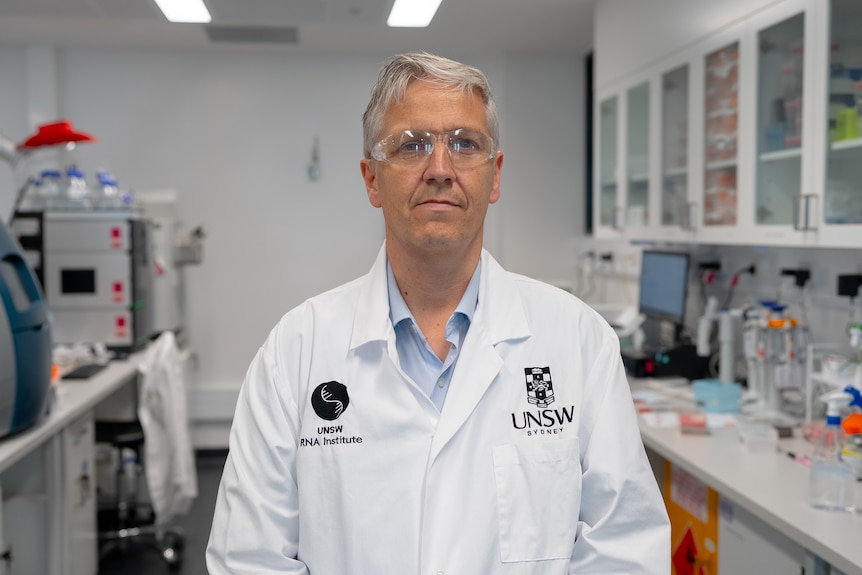 A grey haired man in a lab coat wearing safety glasses.