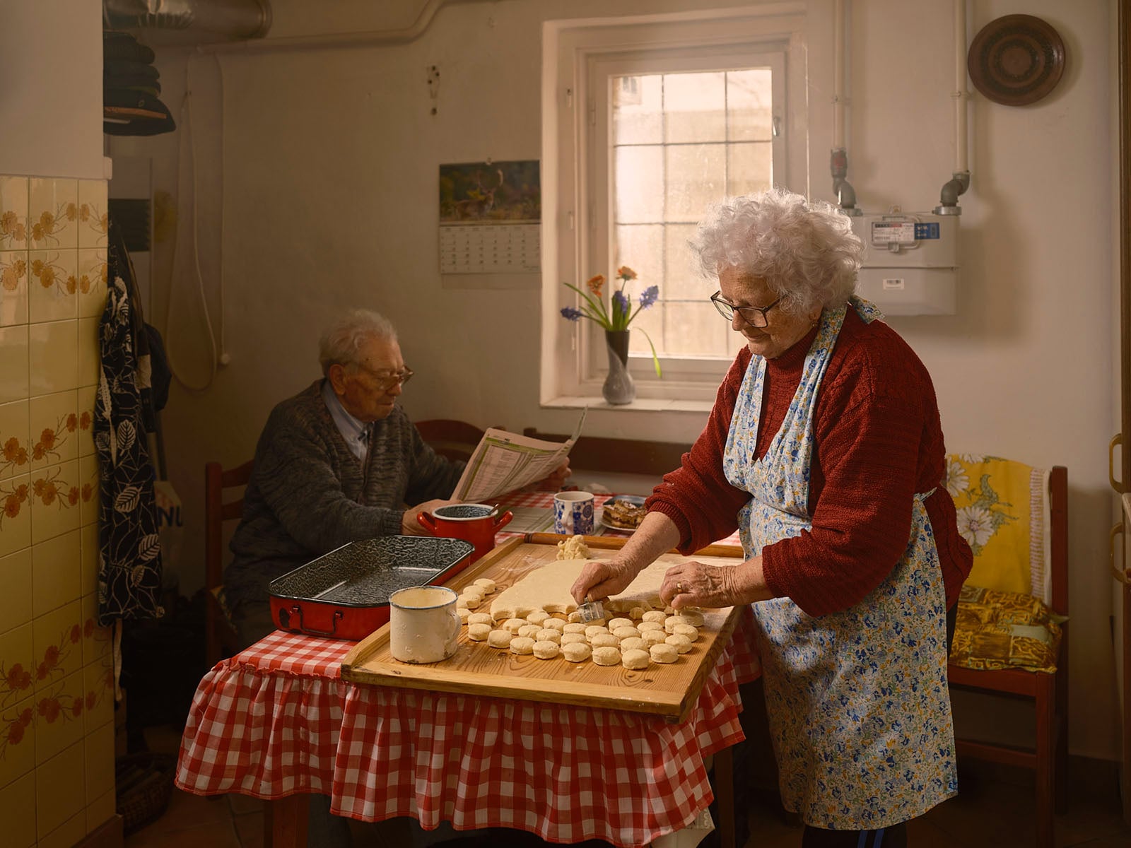 An elderly woman in an apron cuts dough for cookies on a wooden board in a cozy kitchen, while an elderly man sits at a table in the background reading a newspaper. Sunlight streams in through a window with flowers on the sill.