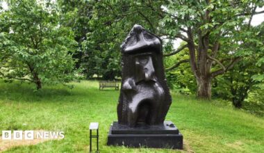 A semi-abstract bronze sculpture on display at a botanic garden. There are trees in the background.