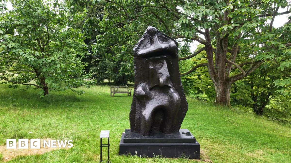 A semi-abstract bronze sculpture on display at a botanic garden. There are trees in the background.