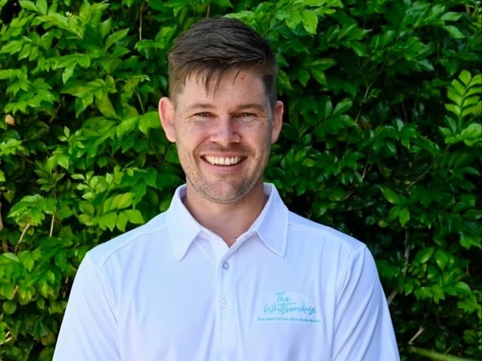 A man smiling at the camera, in front of a hedge.
