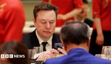 A man in a suit and tie stares at his phone while sitting down opposite a man in a blue jacket in Beijing