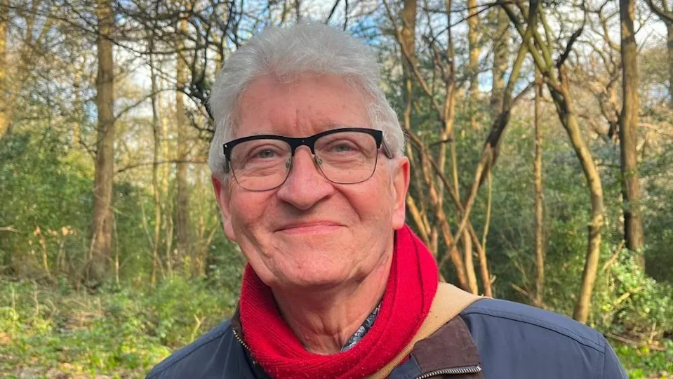 A man with black frame glasses. white hair, a red scarf and a blue coat smiles at the camera. Behind him is a woodland with green foliage and brown, spindly tree trunks.