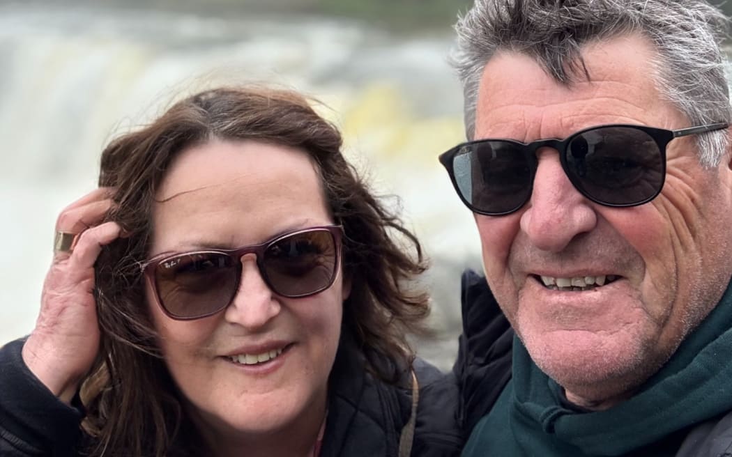This is a photo of Jane and her husband stood in front of Haruru falls. Thy are both wearing sunglasses although it appears to be a bit of a grey day.