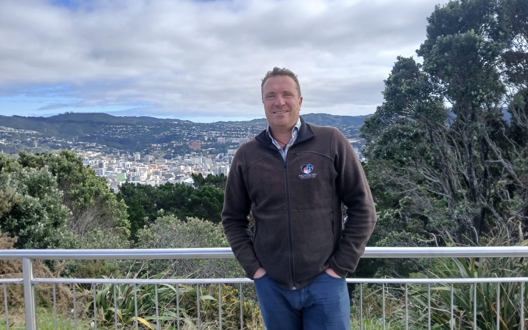 A portrait shot of James Wilcocks at the top of Mount Victoria with a view of some of Wellington city behind him. James is smiling, with his hands in his jean pockets, and is wearing a brown zip jumper.