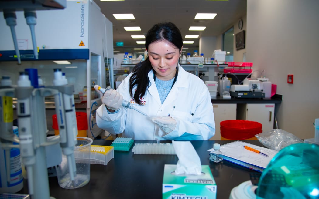 A laboratory researcher uses a pipette to transfer liquid into a small plastic well plate on a bench. The workspace includes racks of tubes, a notebook with a pen, and various lab instruments. Bright overhead lighting illuminates the busy lab environment with equipment and containers arranged behind the work area.