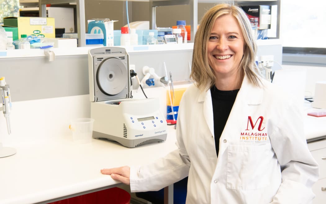 A portrait of a smiling Lisa in a Malaghan Institute white lab coat stands beside a bench with an open centrifuge. The bench holds pipettes, containers, and lab supplies, with shelving and equipment visible in the background.