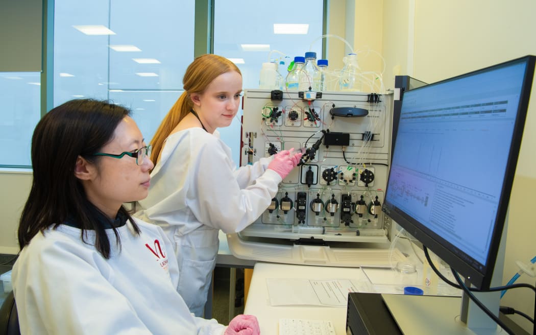 Two laboratory researchers in white lab coats and gloves operate a complex instrument with tubing and control dials connected to bottles. One researcher adjusts the device while another sits at a computer displaying data on a large monitor. The bench includes cables, containers, and lab equipment arranged around the system.