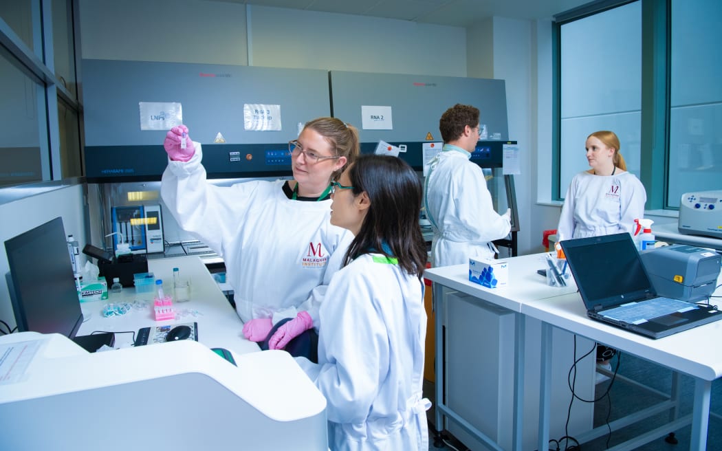 Researchers in a laboratory wearing white lab coats and gloves examine a small vial of liquid. One person holds the vial up to eye level while another looks on. In the background, additional lab workers operate equipment near biosafety cabinets labeled “LNPs” and “RNA 2,” with benches, instruments, and pipettes visible throughout the room.