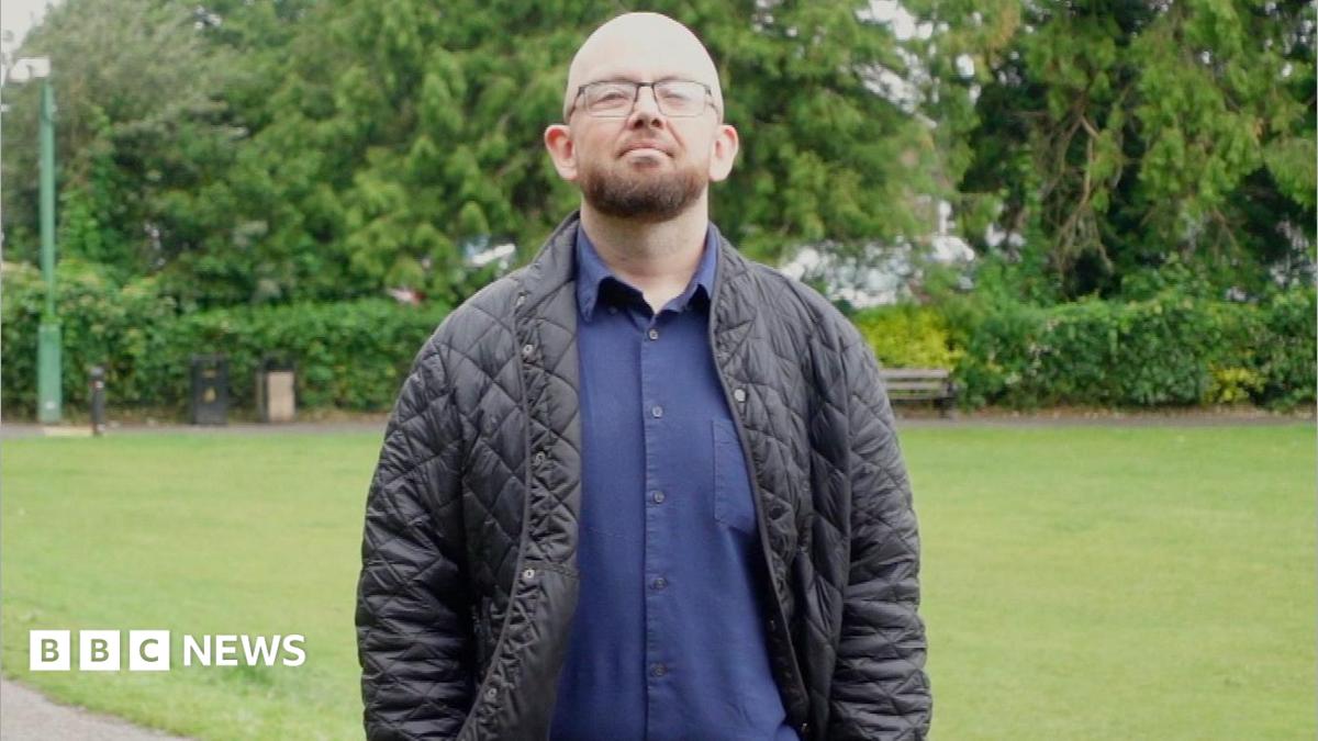 A bald and beared bespectacled man in his thirties, wearing a blue shirt and trousers and a dark jacket, stands in a park with his hands in his pockets.