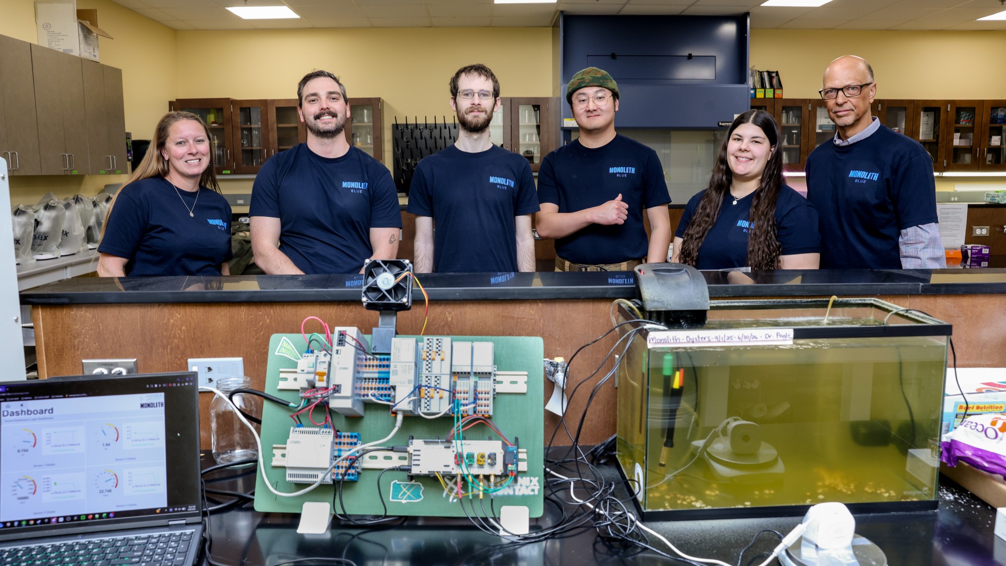 six people in matching t-shirts stand in a classroom behind a tank of turbid water containing small mollusk shells with wires coming out of the tank connected to a row of electronics and computers