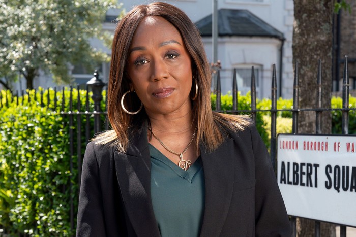 Diane Parish as Denise Fox, with her arms folded stood in front of the Albert Square sign in key art for EastEnders