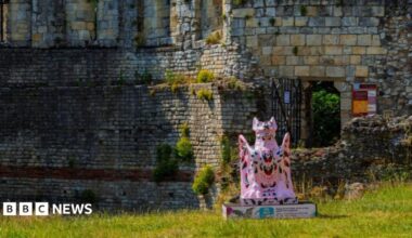 A pink-coloured sculpture of a grotesque creature placed near ruined walls. It sits