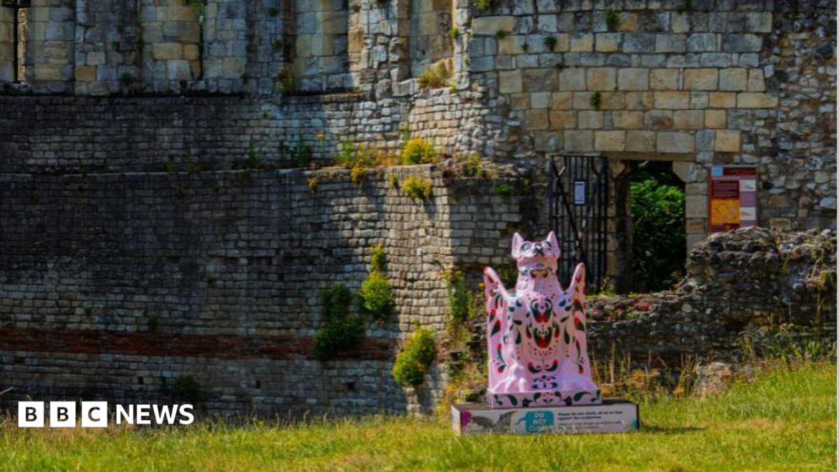 A pink-coloured sculpture of a grotesque creature placed near ruined walls. It sits