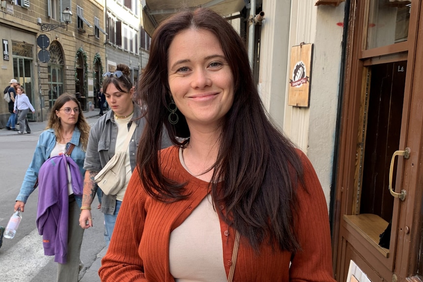Image of hannah who has long hair and smiles at camera. Wears a cardigan and top. Two people walk behind her