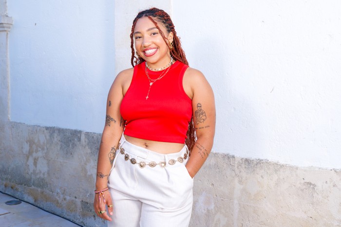 A woman stood smiling in front of a white background, wearing a red crop top and white trousers.