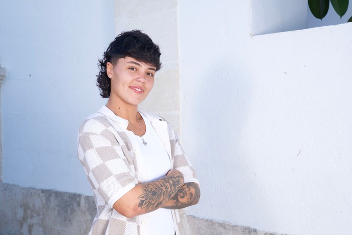 A woman stood in front of a white wall smiling ahead with her arms folded in front of her chest.
