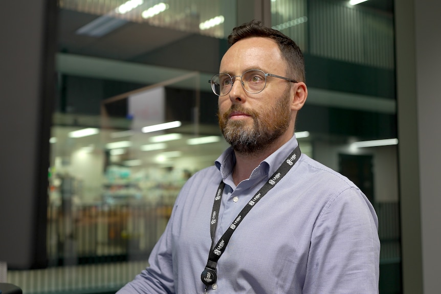 Martin Smith in a blue long sleeve and lanyard, glasses, sitting at a desk and looking at a laptop monitor.