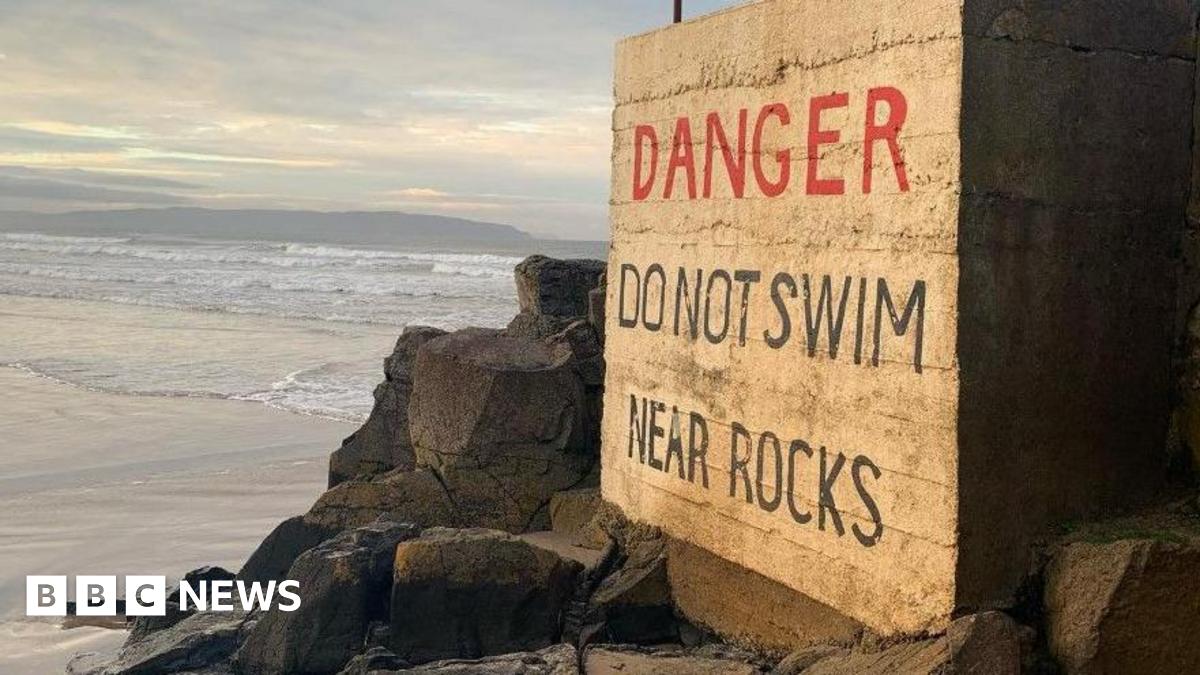 Portstewart Strand. A large sign reads: 'Danger – Do Not Swim Near Rocks.' It is positioned beside a wide area of jagged black rocks, with the shoreline visible in the background.