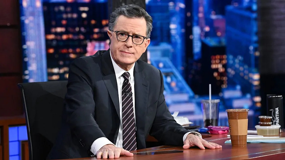 Stephen Colbert sits with a tea cup at his desk staring at the camera during a final season episode of The Late Show. He has glasses and suit on.