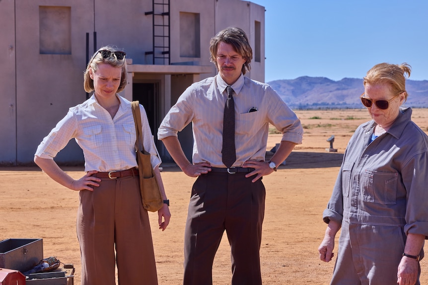 Three people stand outside in a dusty flat area.